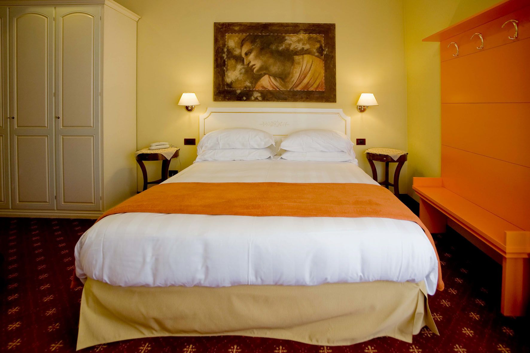 A hotel room with a large bed, two bedside tables, a nightstand, a wardrobe, and a painting on the wall.