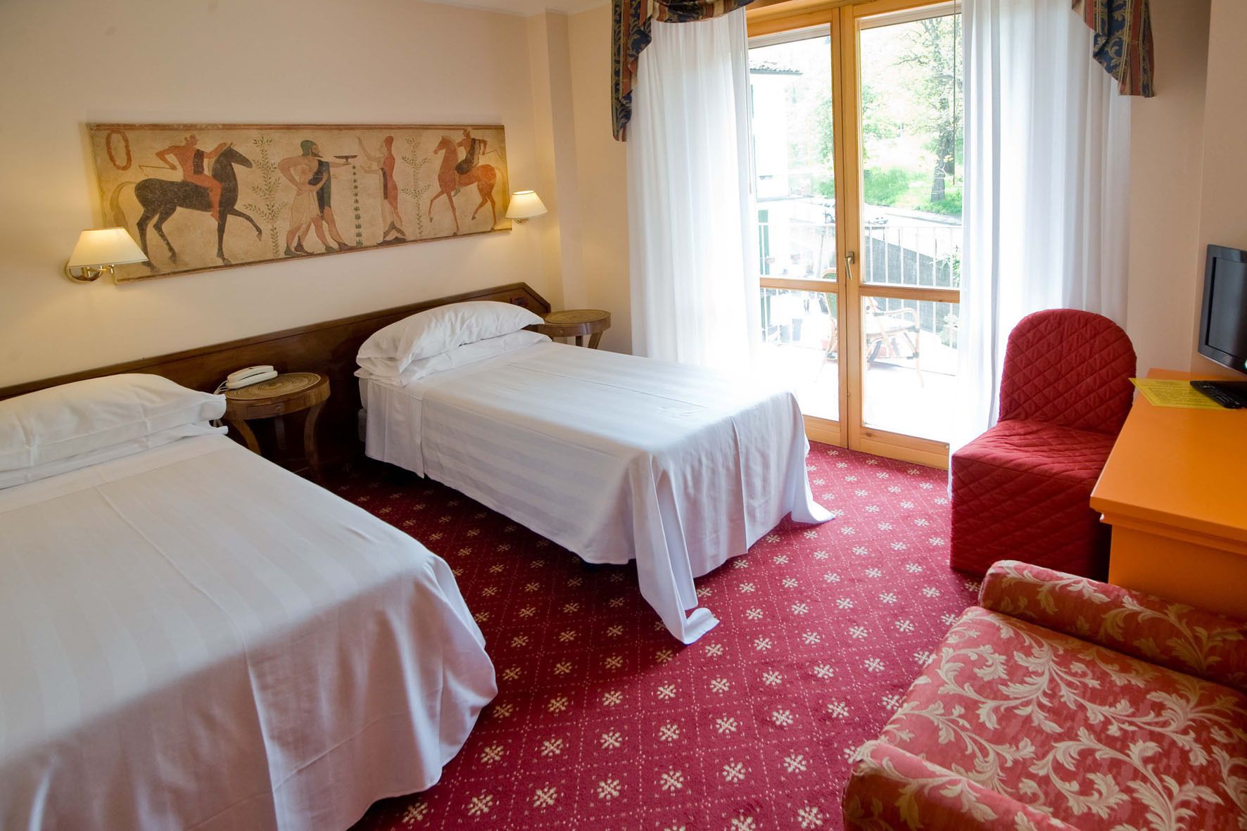 Habitación de hotel con dos camas individuales, un sillón rojo, un televisor y un mural en la pared.