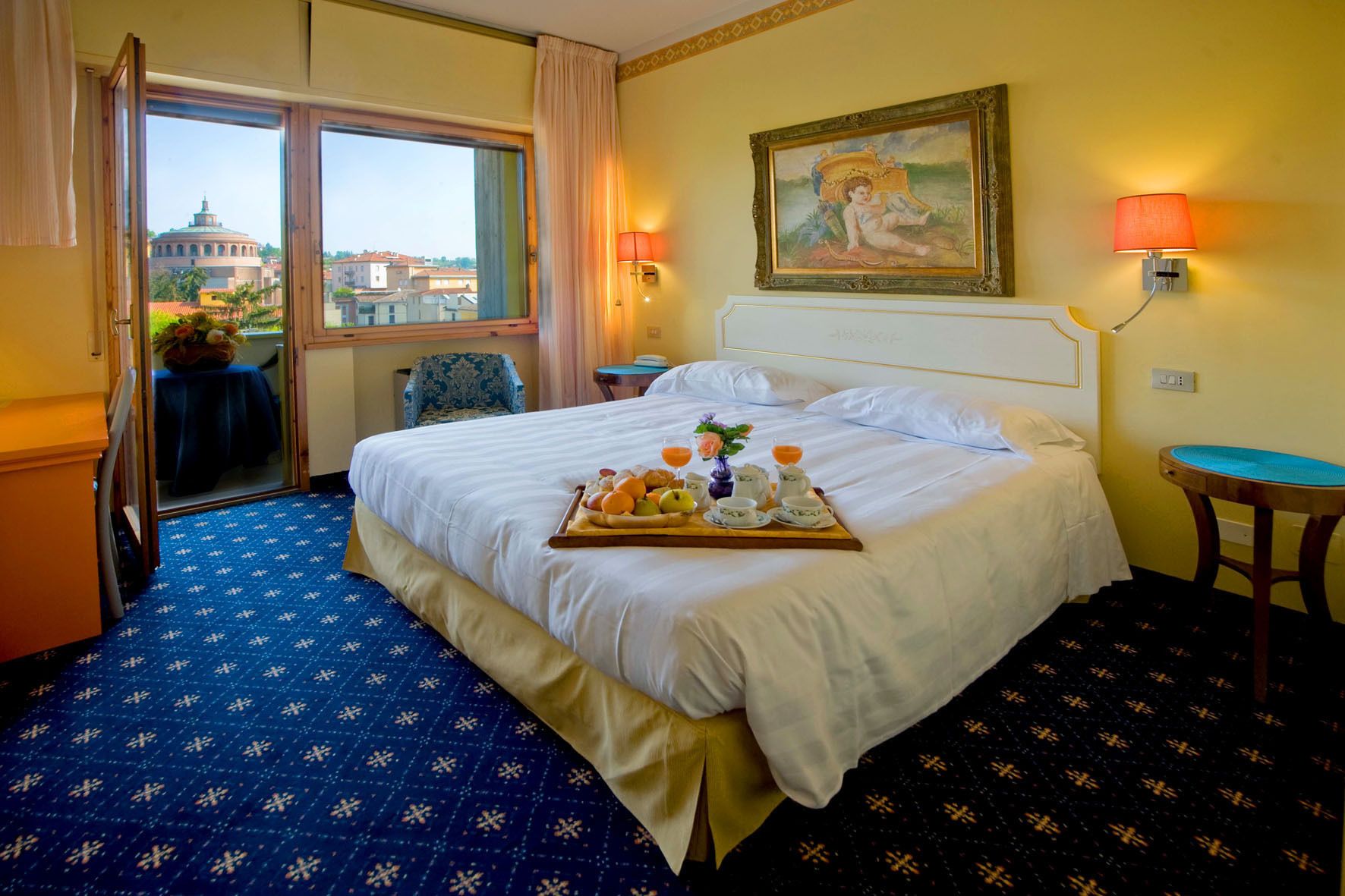 A hotel room with a bed, a breakfast tray, a painting, and a view of a building through the window.