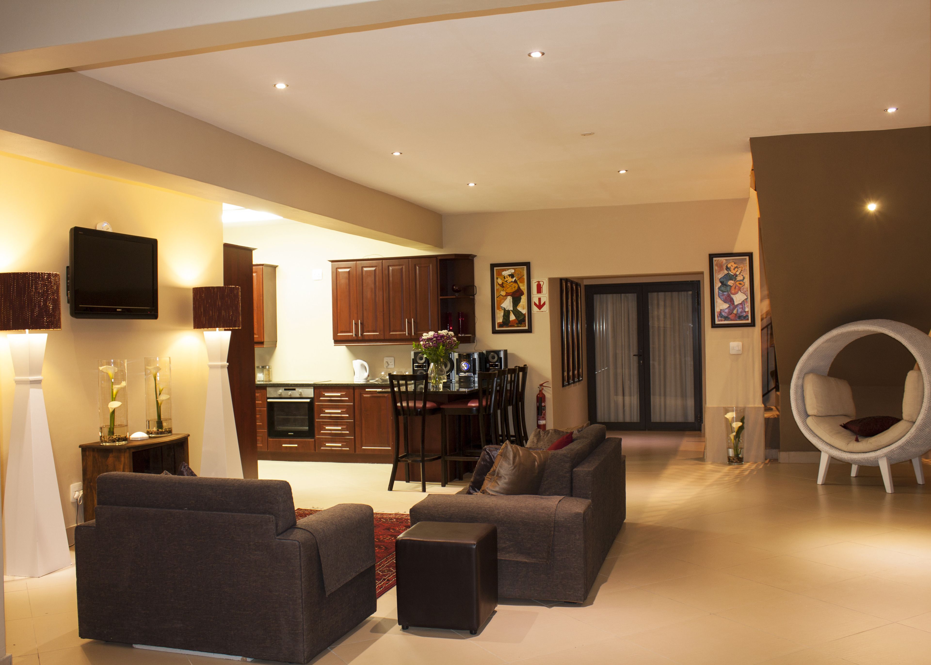 A modern living room with a sofa set, a dining area with a table and chairs, a kitchen with wooden cabinets, and a large window with curtains.