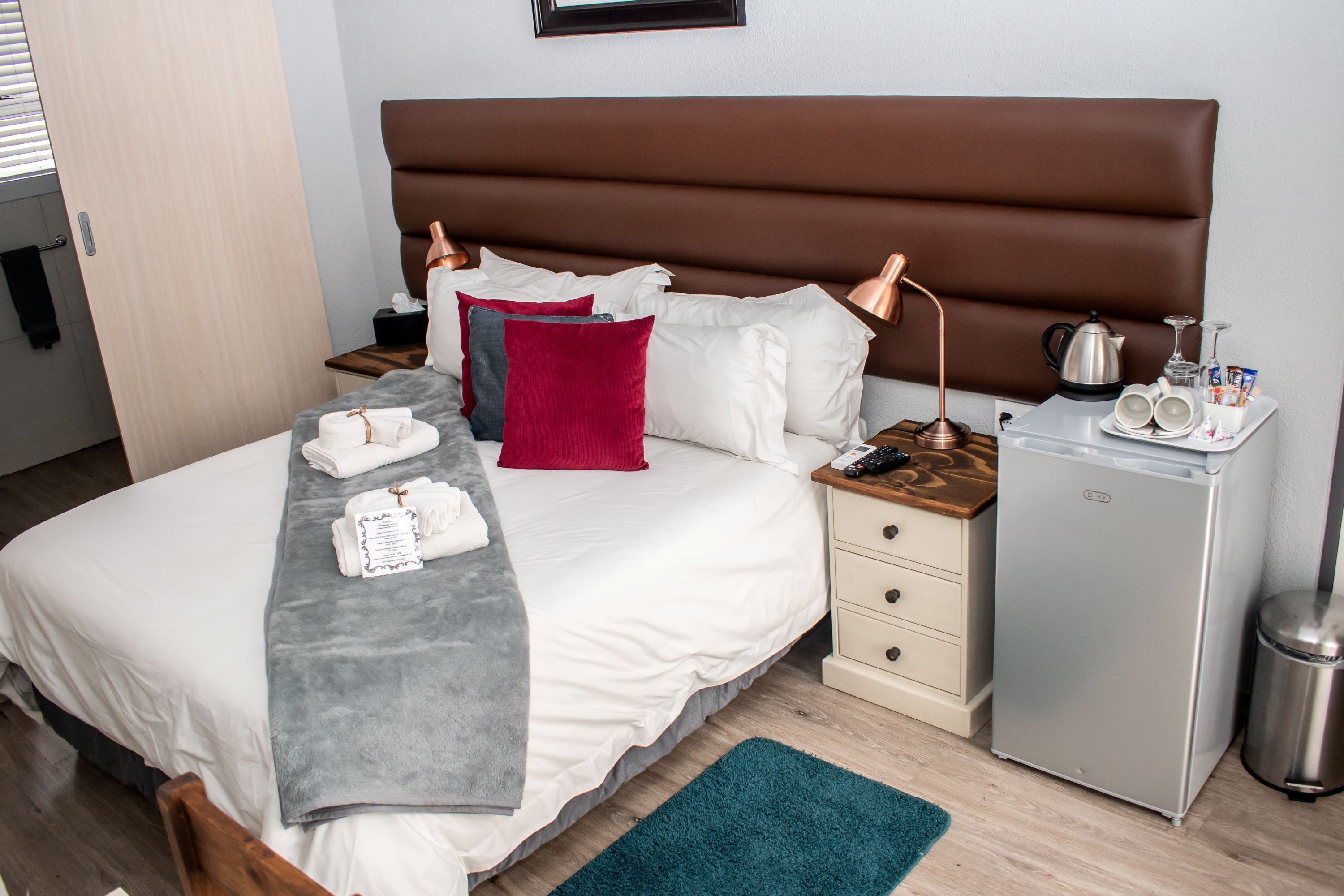 A neatly made bed with white linens, a brown headboard, and a teal rug. There is a small bedside table with a lamp, a mini fridge, and a trash can.