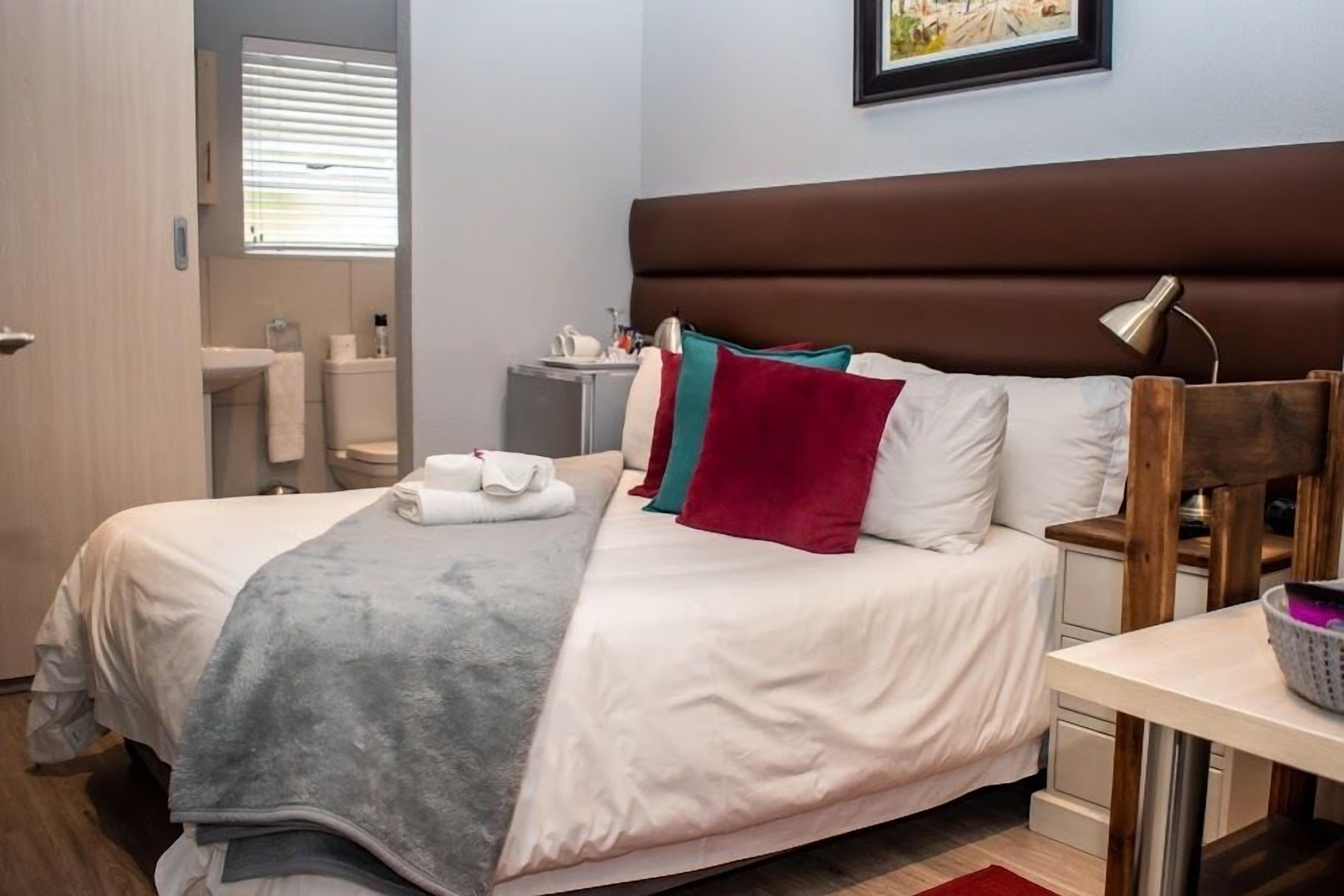 A neatly made bed with a grey blanket, a brown headboard, and colorful pillows. There is a small bedside table with a lamp and a basket.