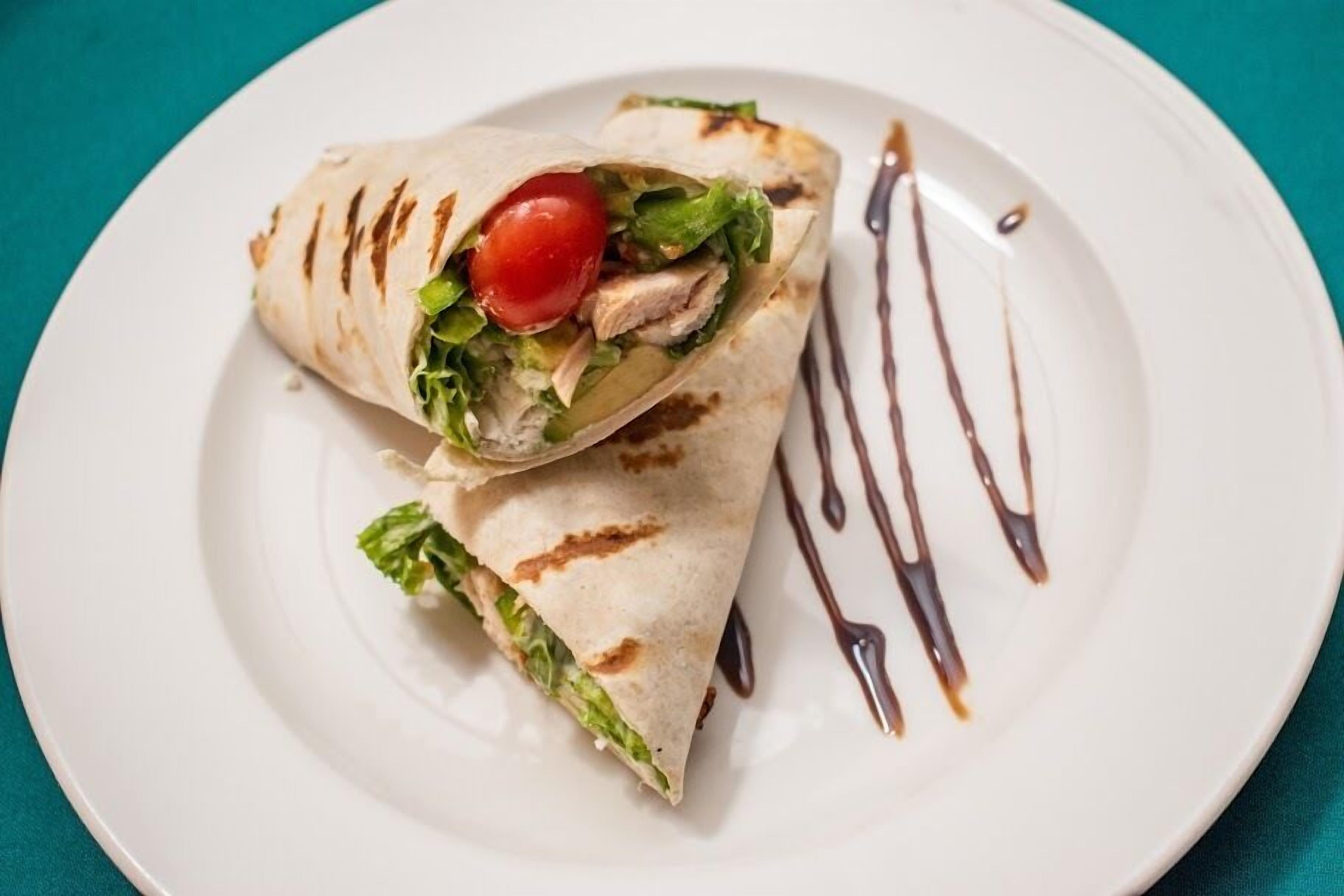 A wrap with lettuce, tomato, and chicken on a white plate with a drizzle of sauce.