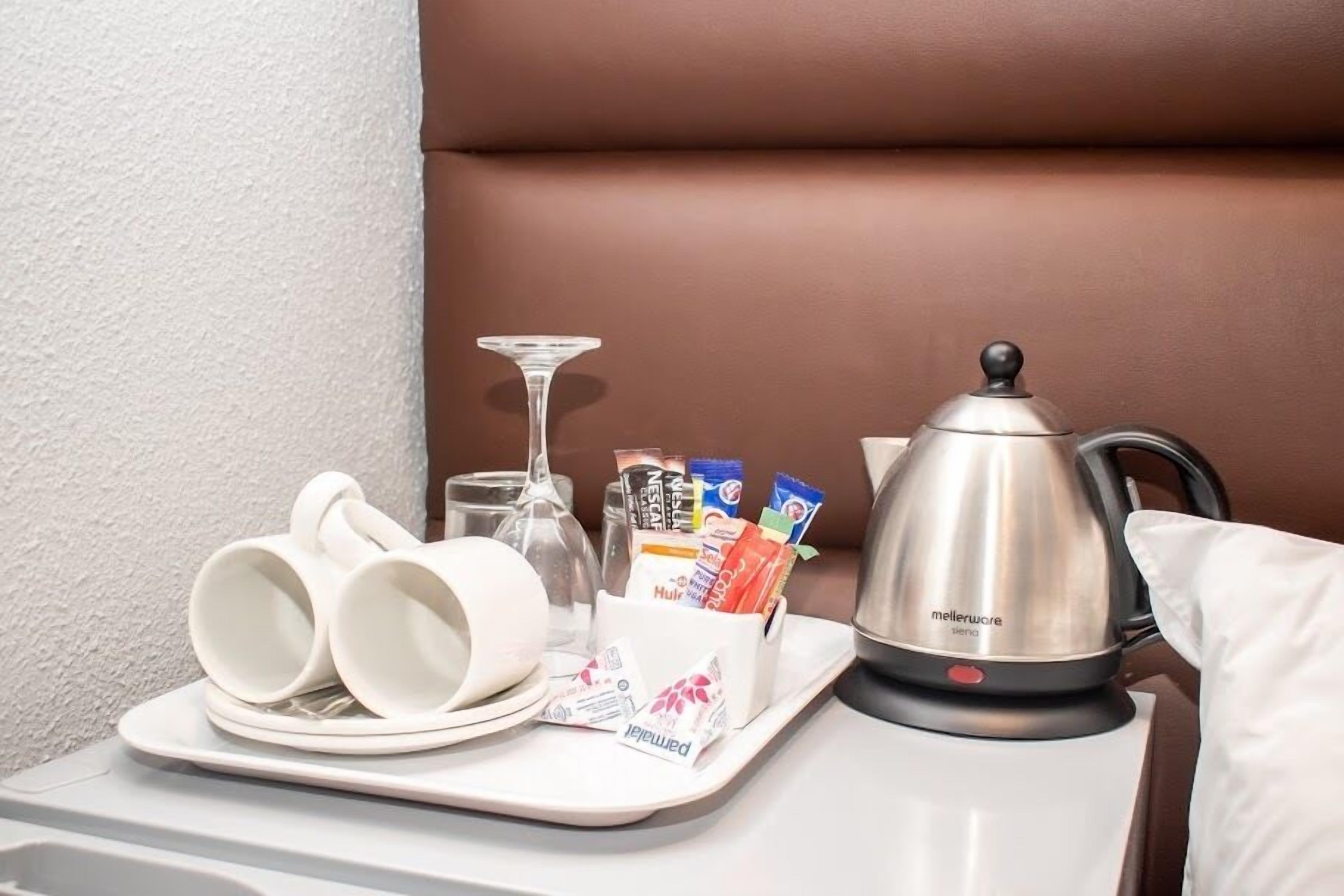 A tray with white cups, a glass, a coffee pot, and a container with snacks on a table.