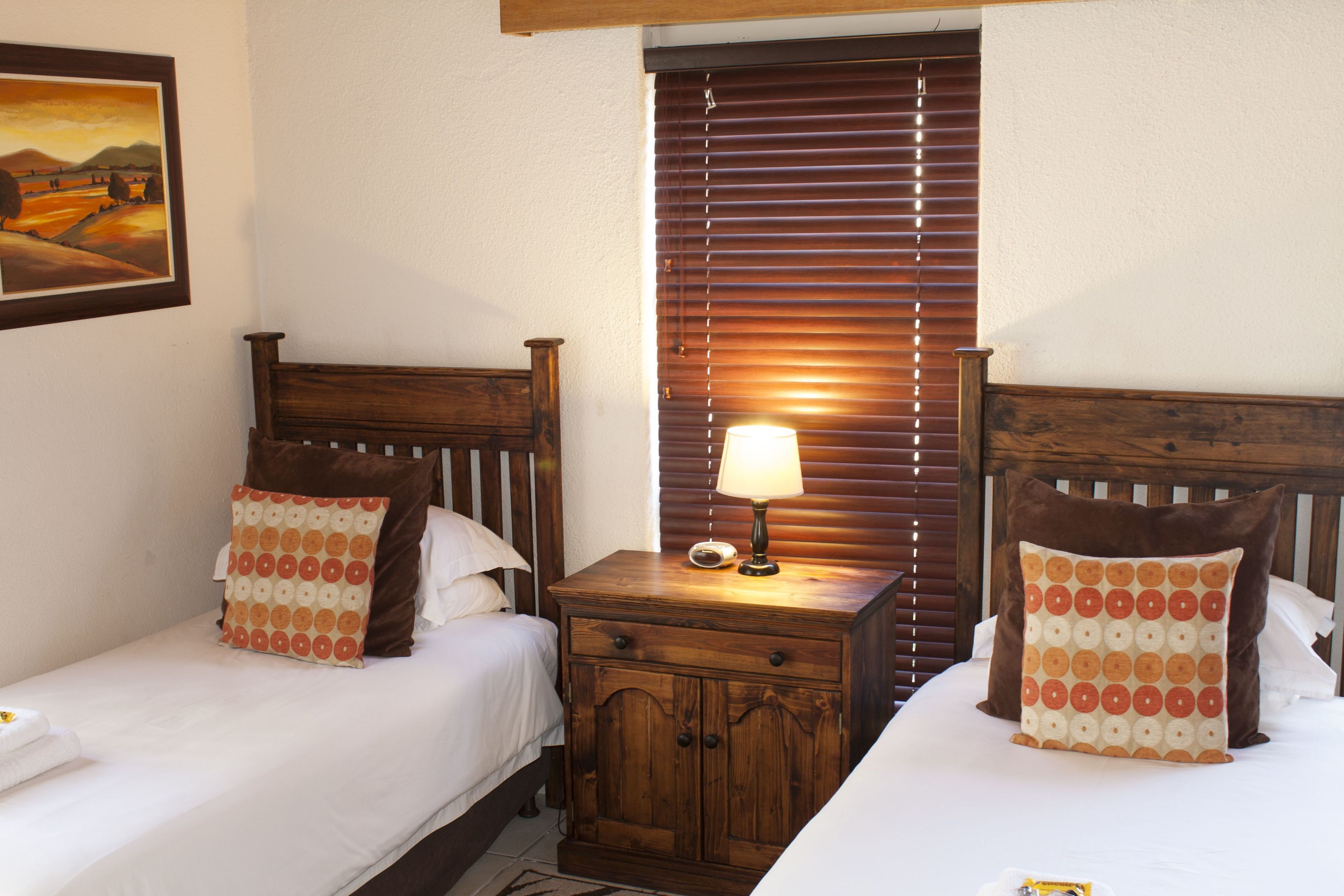 A hotel room with two beds, wooden headboards, a nightstand with a lamp, and a painting on the wall.