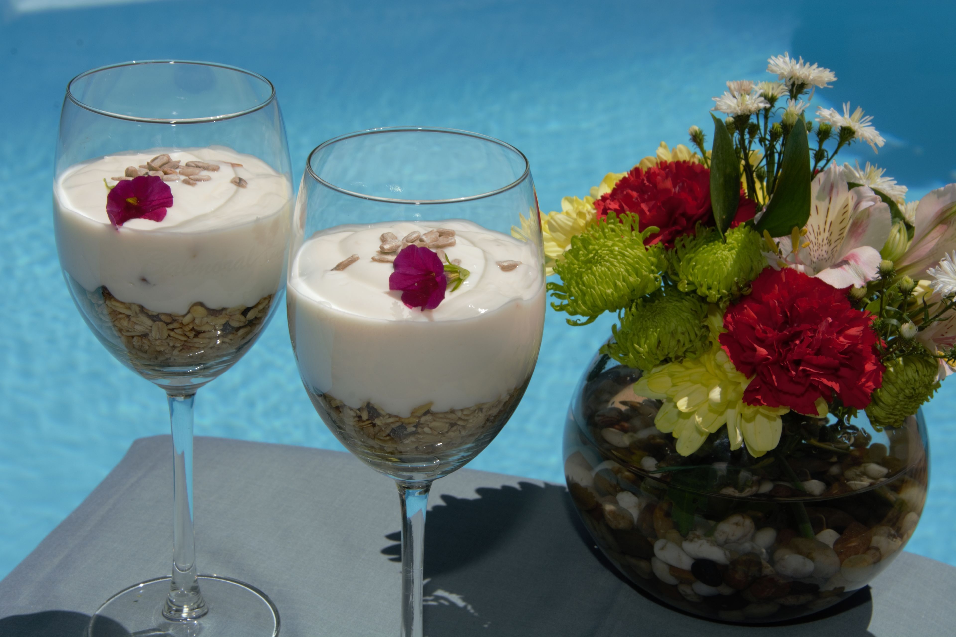Two glasses of yogurt parfait with granola and a purple flower on top, placed on a table next to a vase with colorful flowers.