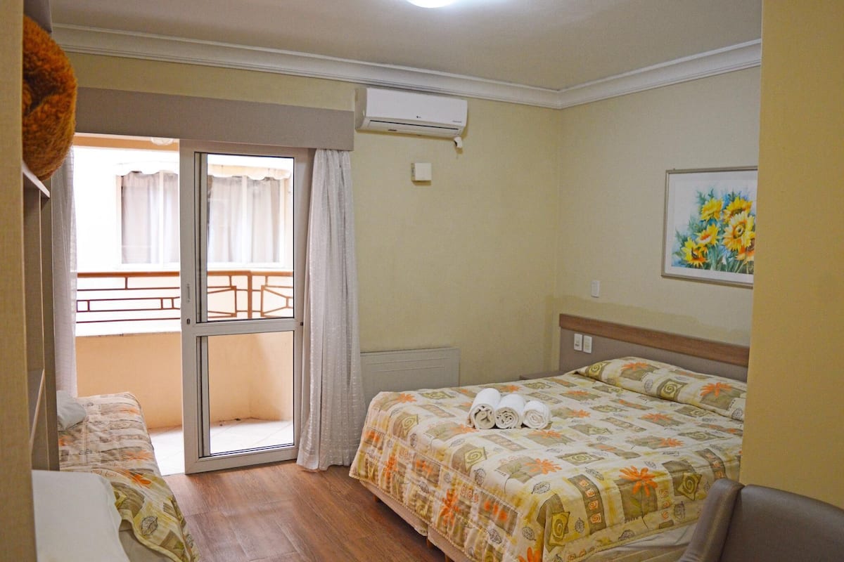A hotel room with a bed, a chair, a desk, a window with curtains, an air conditioning unit, and a painting on the wall.