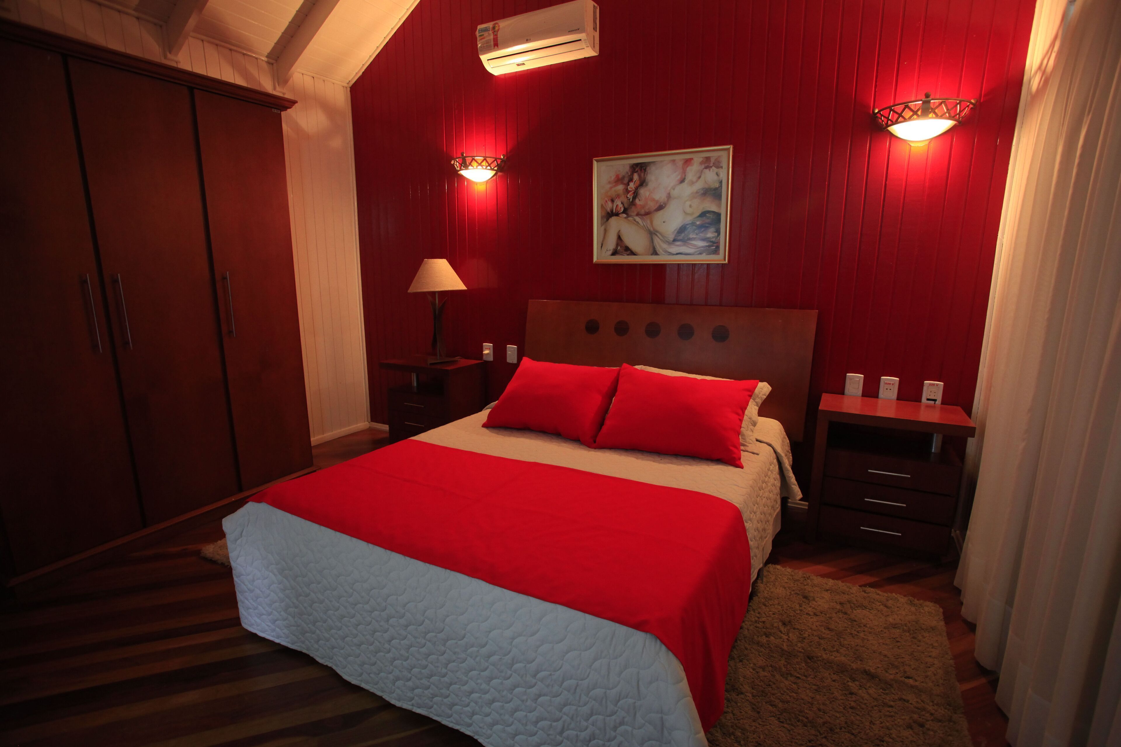 A bedroom with a bed, red walls, a painting, and a nightstand with a lamp.