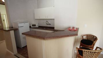 A compact kitchen with a refrigerator, microwave, and a small sink. There are two wicker chairs and a small table with a red object on it.