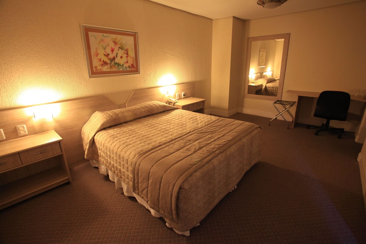 Une chambre d’hôtel avec un lit, des lampes de chevet, un bureau avec une chaise, un miroir et un tableau au mur.
