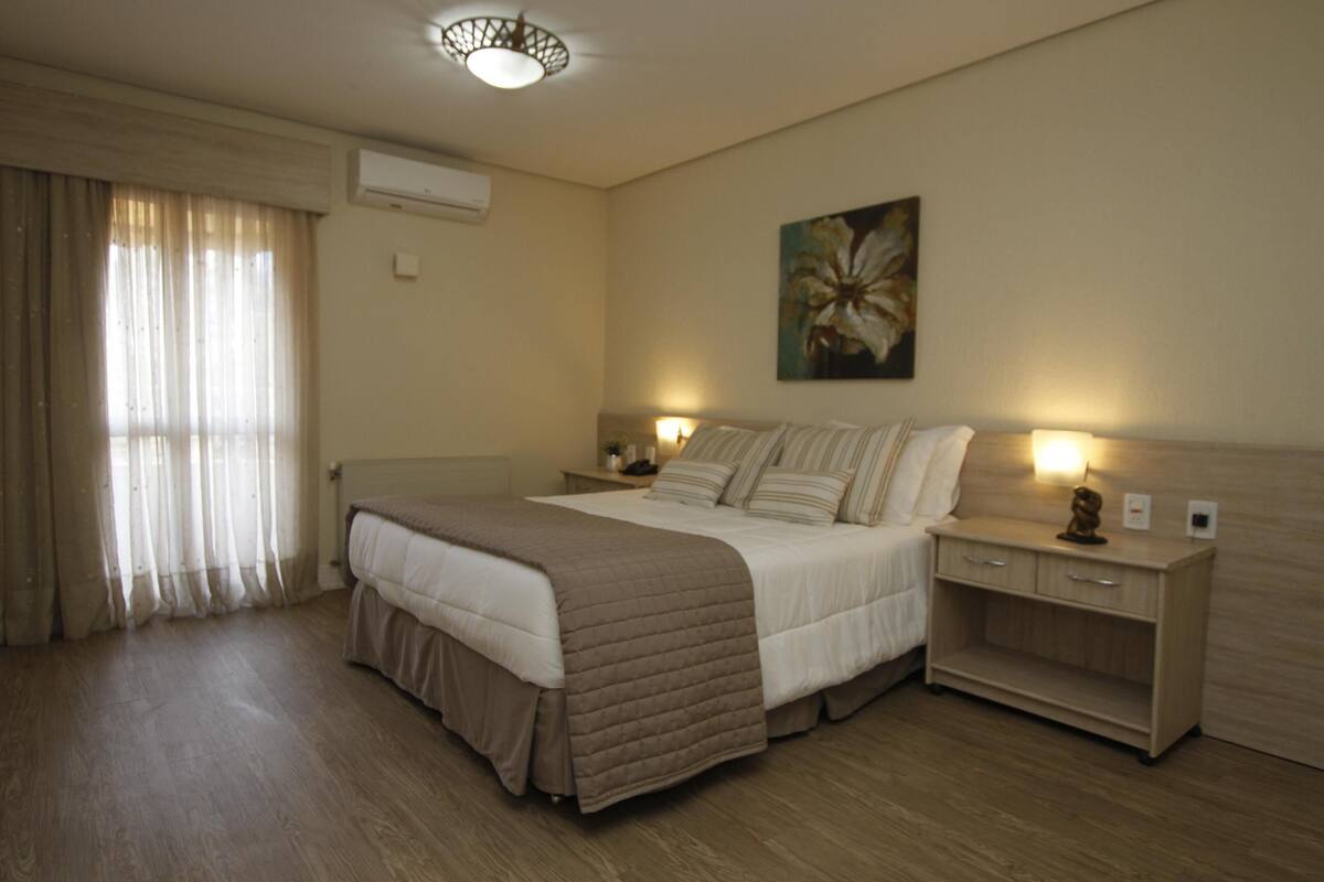 A hotel room with a bed, bedside tables, a nightstand, a lamp, a painting on the wall, and a window with curtains.