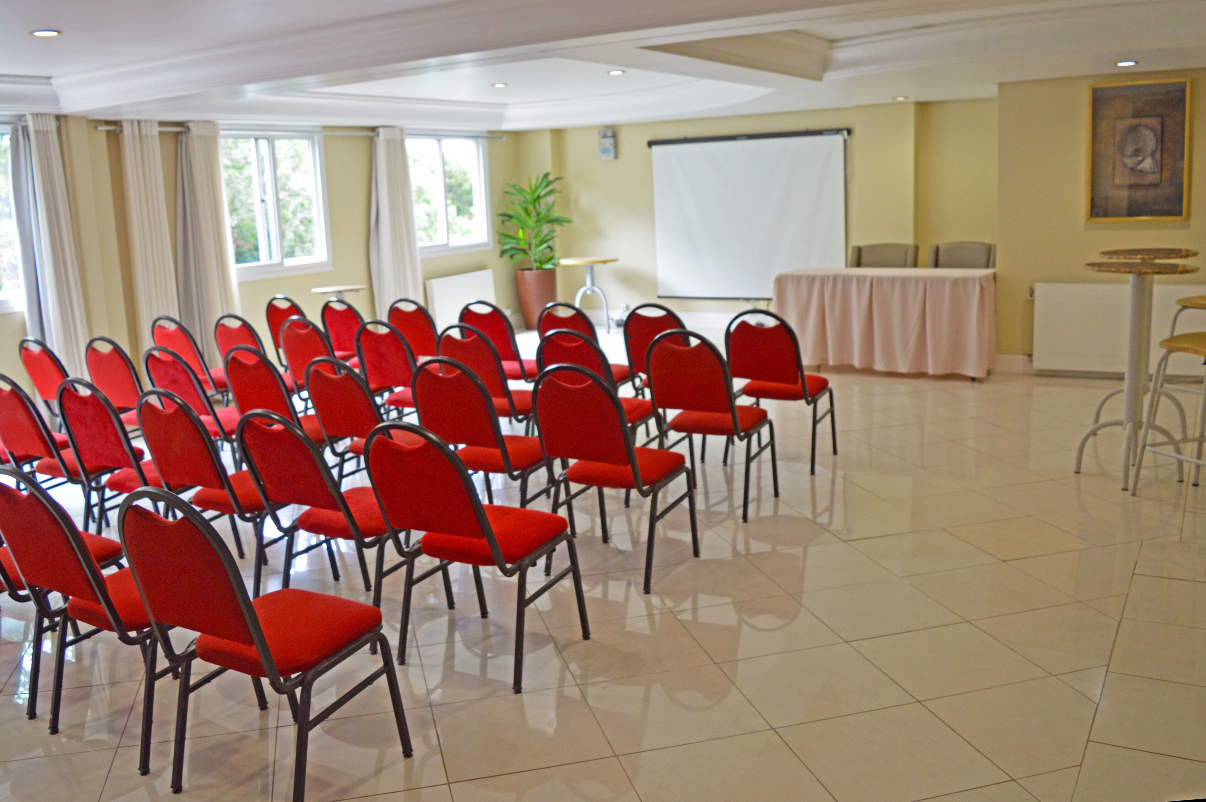 A conference room with rows of red chairs, a projector screen, a table with a tablecloth, and a potted plant.