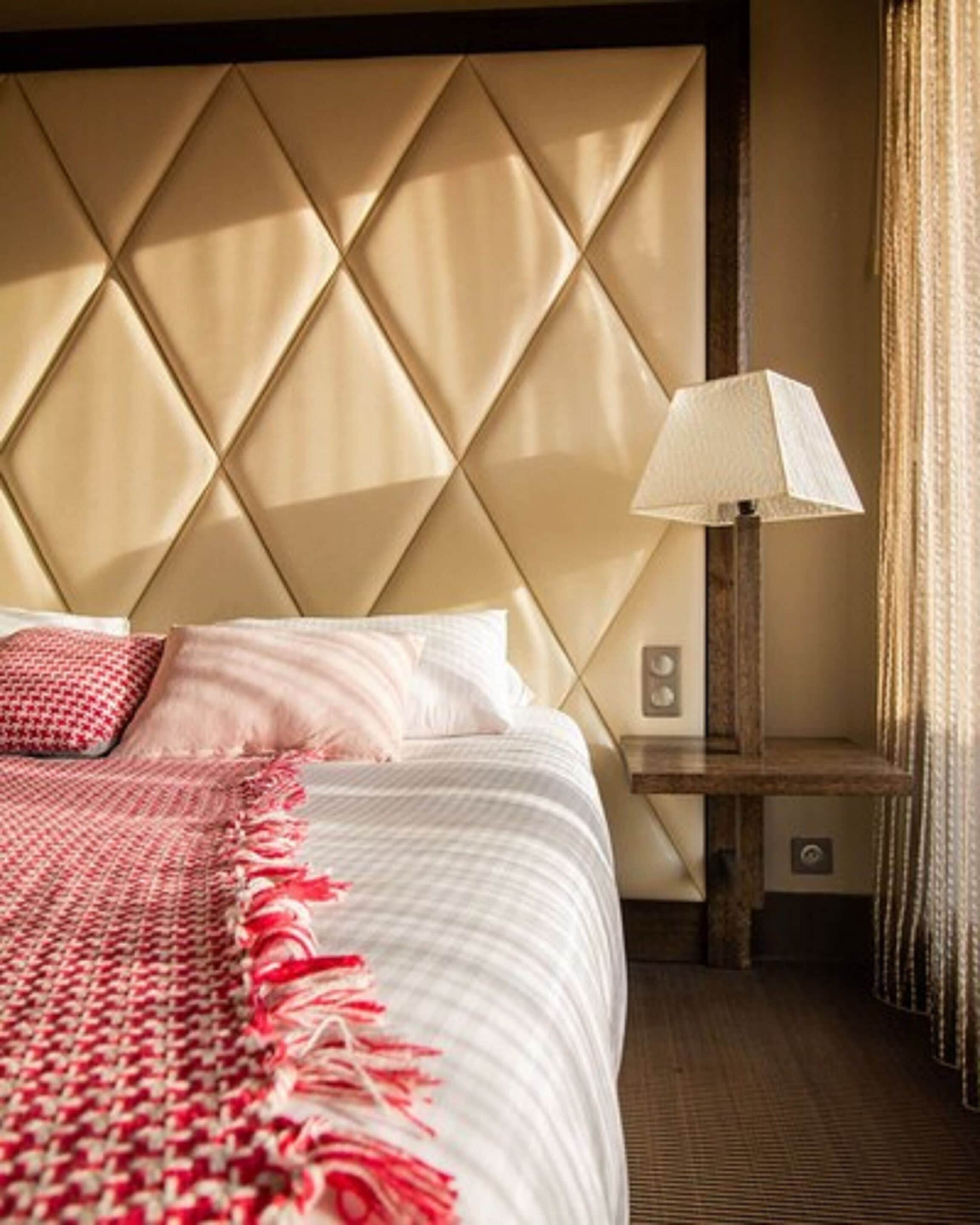 A hotel room with a large bed, a headboard with a diamond pattern, a bedside table with a lamp, and a red and white striped throw blanket.