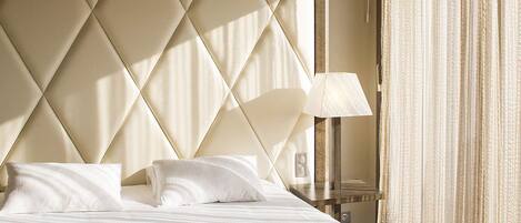 A hotel room with a large bed, a headboard with a diamond pattern, a bedside table with a lamp, and a window with curtains.
