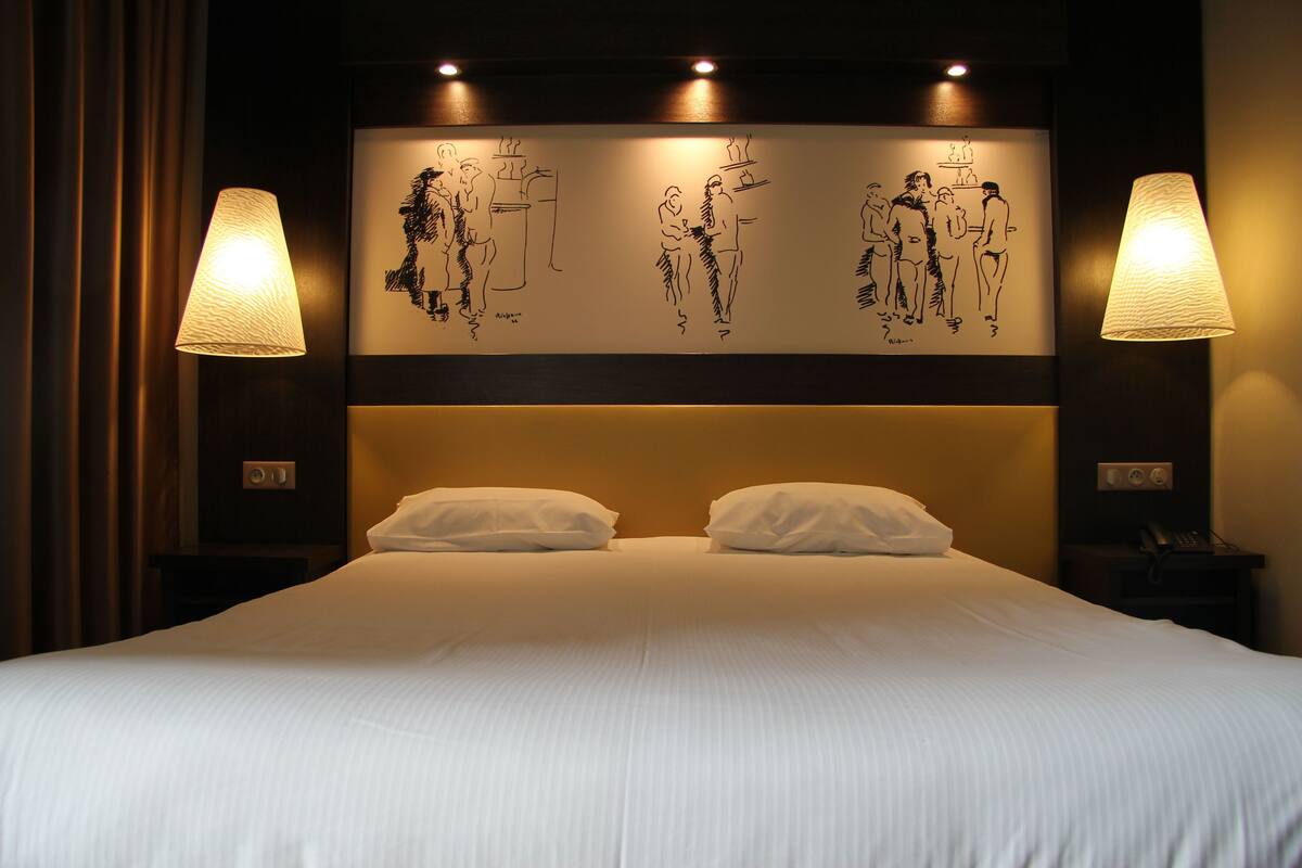 A hotel room with a large bed, two bedside lamps, and a wall with black-and-white sketches.