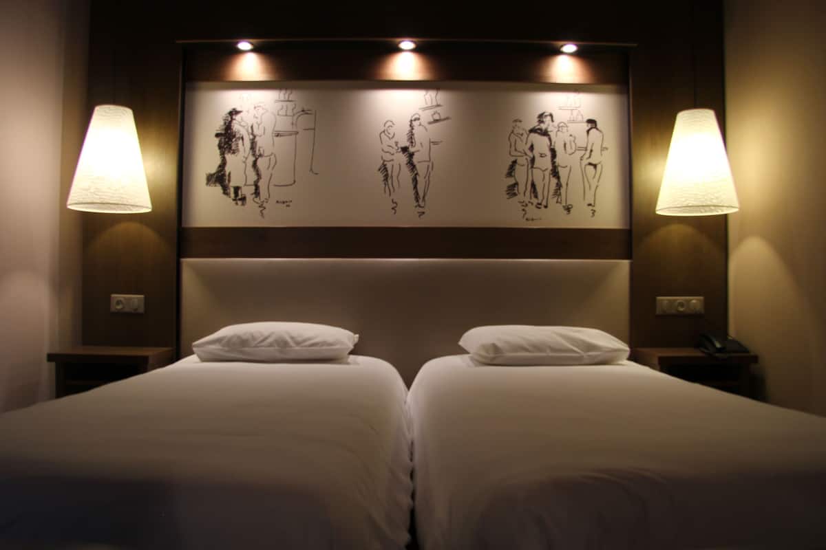 A hotel room with two beds, a headboard with wall art, and bedside lamps.
