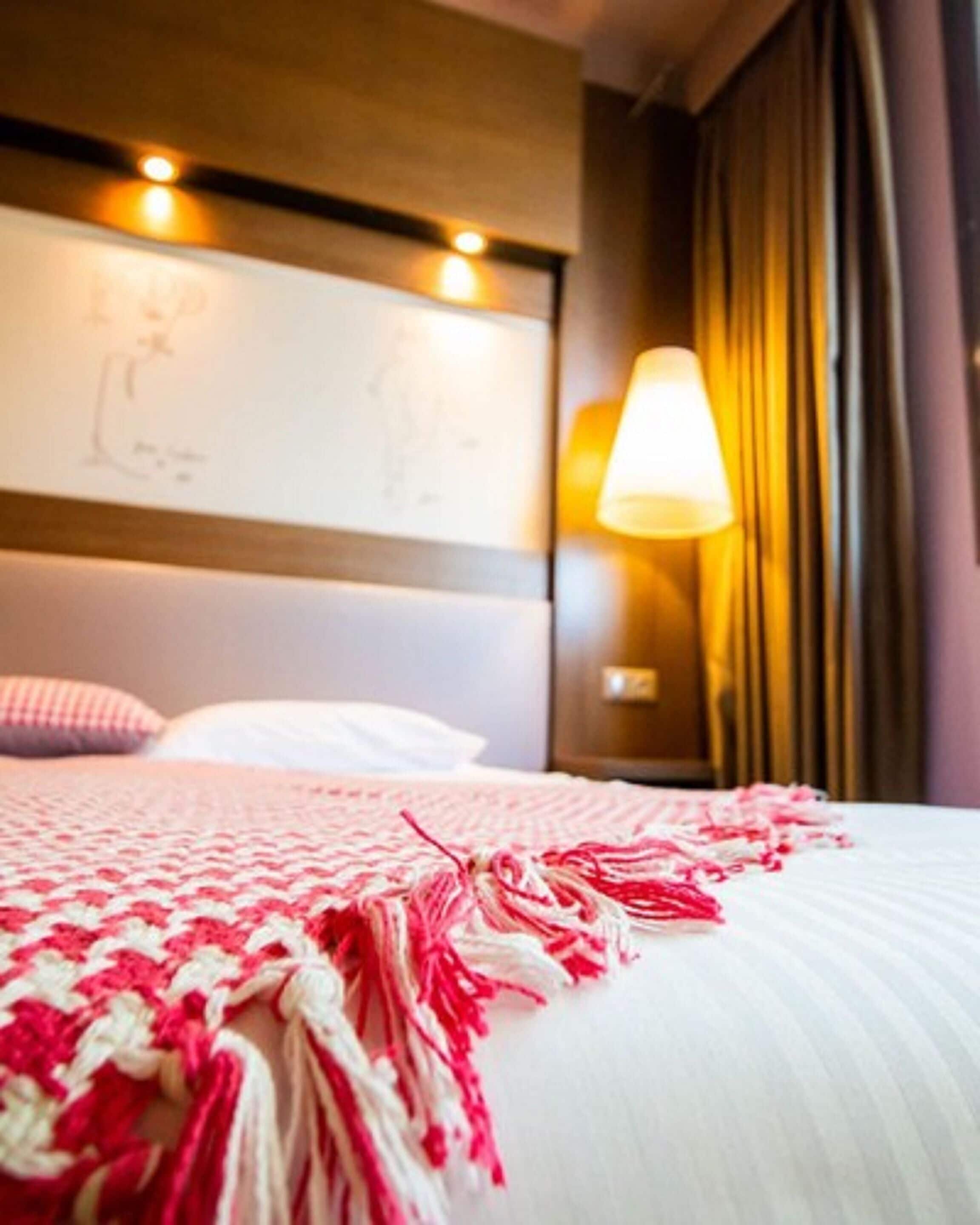 A hotel room with a bed, a headboard with a light fixture, a bedside lamp, and a window with curtains.