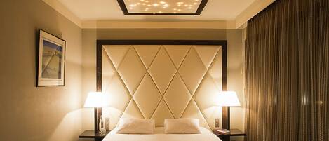 A hotel room with a large bed, two bedside lamps, a diamond-patterned headboard, a framed picture on the wall, and a curtain on the window.