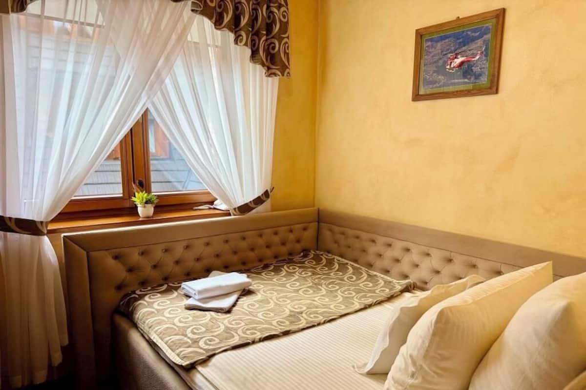 A hotel room with a bed, a window with curtains, a small potted plant, and a painting on the wall.