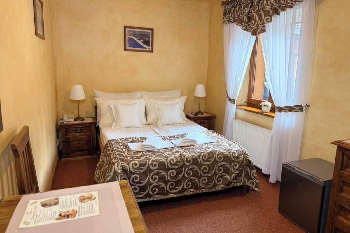 A hotel room with a bed, bedside tables, a small refrigerator, and a window with curtains.