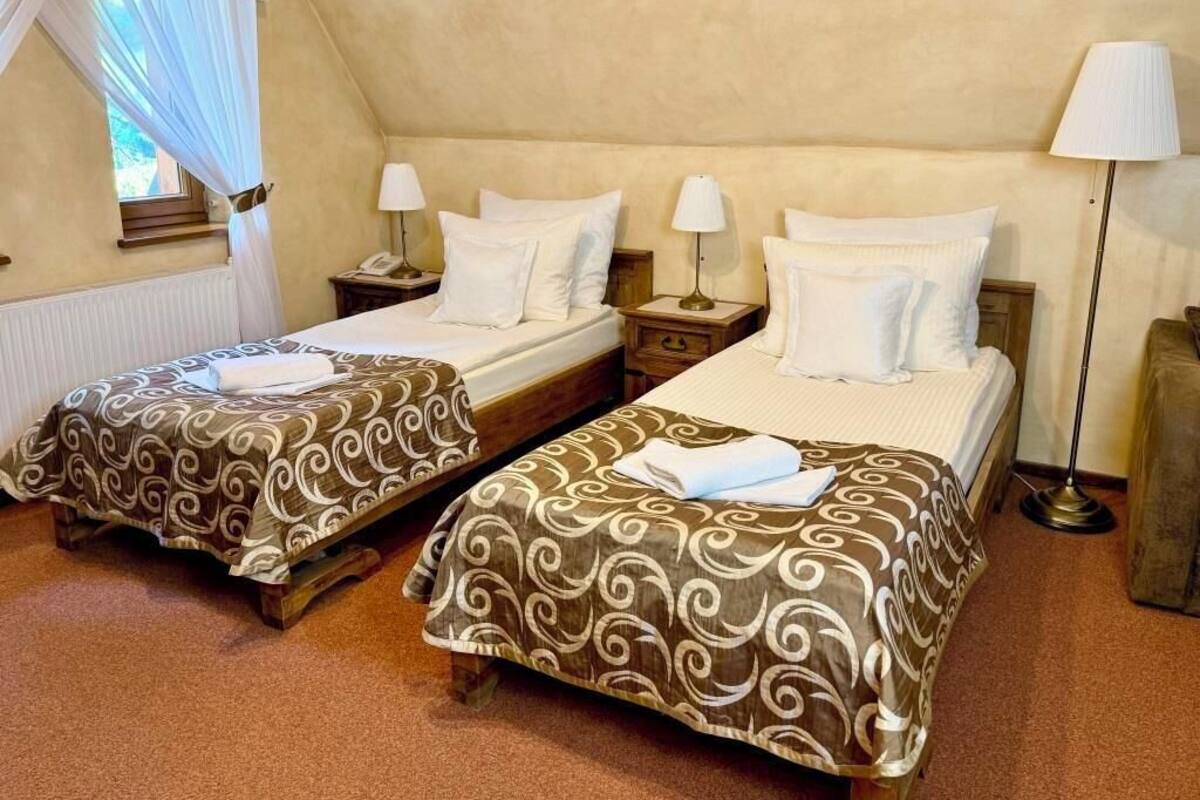 A room with two beds, each with a patterned bedspread and white pillows, a bedside table with a lamp, and a window with curtains.
