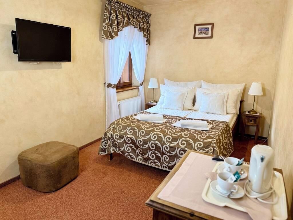 Quarto de hotel com cama, televisão, mesa de cabeceira com luminária, um banquinho e um suporte com xícaras e chaleira.