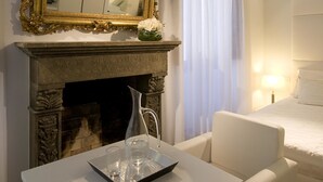 Een slaapkamer met een open haard, een wit bed, een witte fauteuil, een tafel met dienblad en een grote, versierde spiegel.