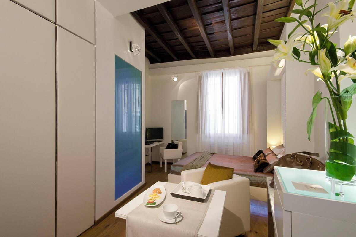 Una habitación de hotel moderna con una cama, un área de descanso con una mesa dispuesta para el desayuno, un televisor y un gran ventanal con cortinas.