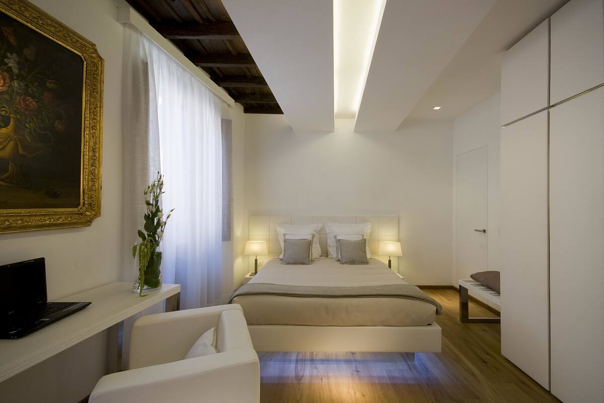 Un dormitorio moderno con cama, mesitas de noche, un escritorio con computadora portátil, una silla y un cuadro grande enmarcado.