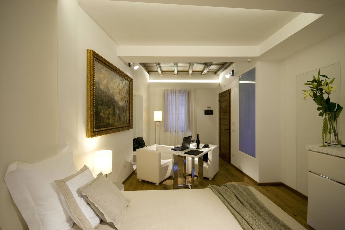 Habitación de hotel con una cama, un escritorio con una computadora portátil, una silla, una lámpara y un cuadro en la pared.