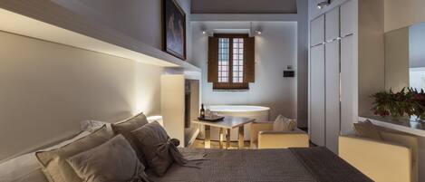 A bedroom with a bed, a bedside table, a window, a mirror, and a small sitting area with a table and chair.