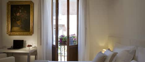 A hotel room with a bed, a desk with a laptop, a painting, and a view of a balcony with flowers.