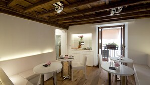 Een gezellige kamer met houten plafond, witte wanden en een balkon met potplanten. Er staan twee ronde tafels met stoelen, een witte bank en een klein keukentje met een presentatie van fruit.