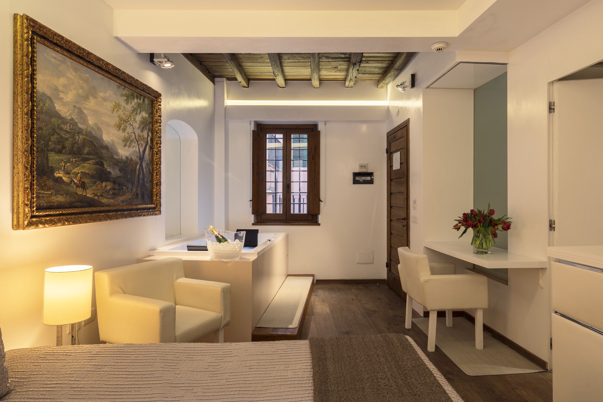 A hotel room with a bed, a desk with a chair, a large painting, a vase with flowers, and a window with blinds.