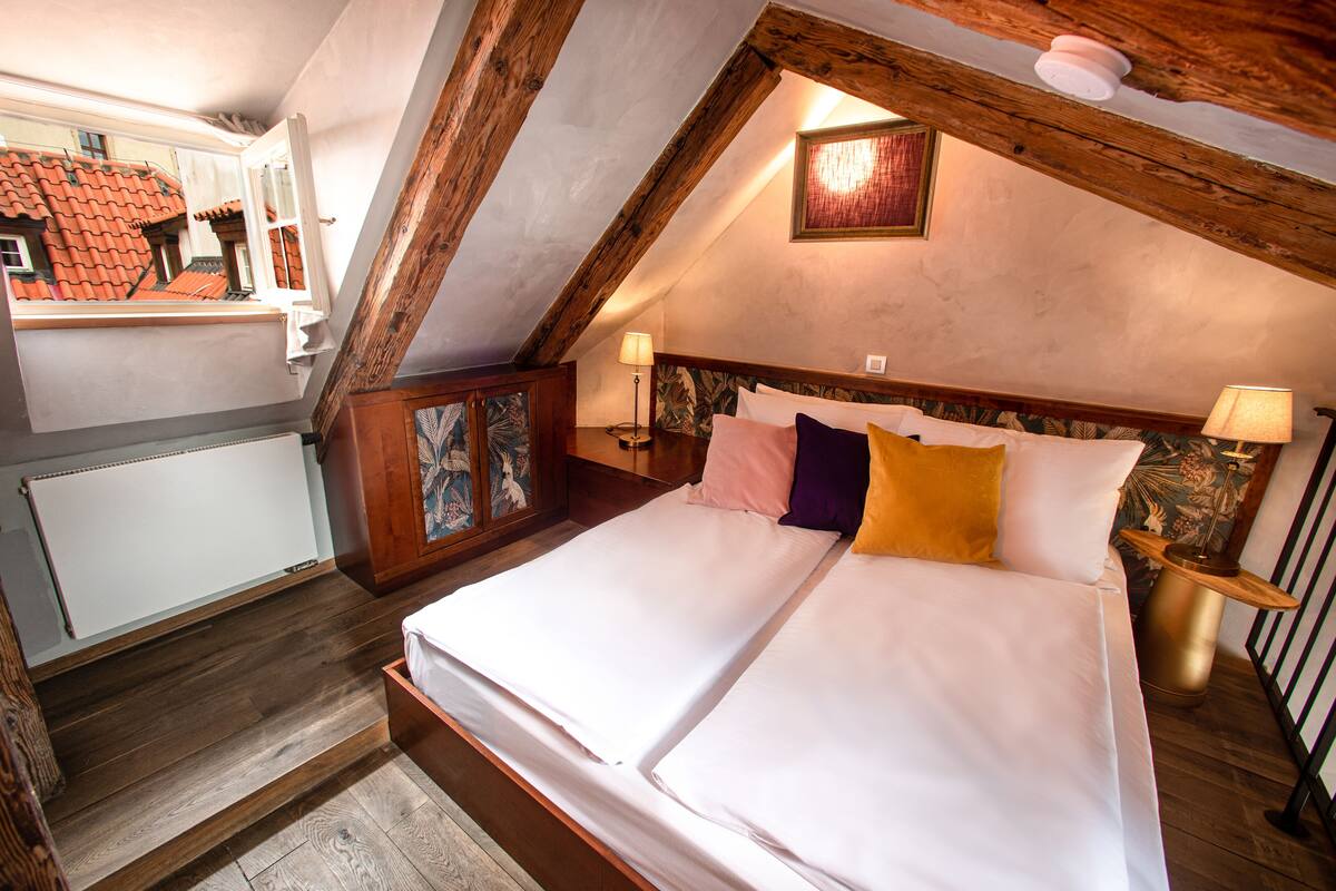 A bedroom with a large bed, wooden beams, a radiator, a wooden cabinet with artwork, and a window with a view of buildings.