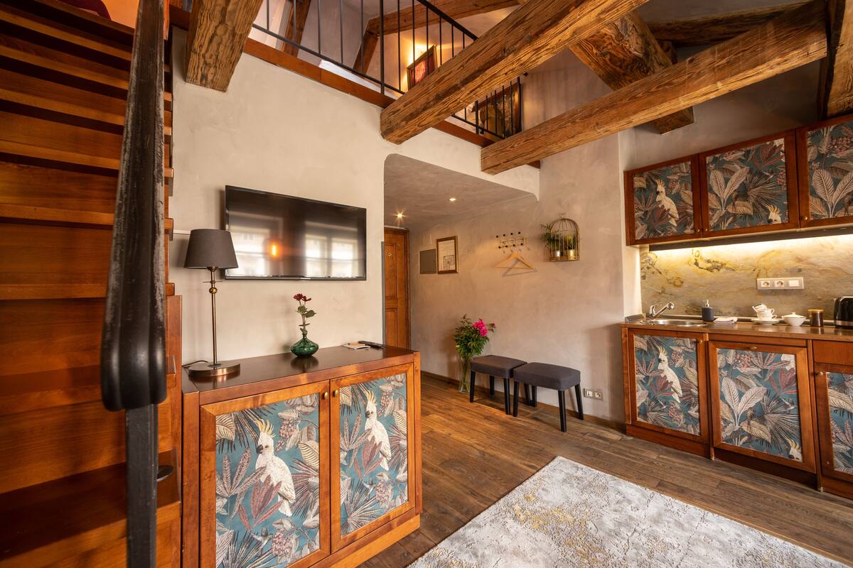 A spacious living area with wooden flooring, a fireplace, a flat-screen TV, and a kitchen with decorative leaf-patterned cabinets.