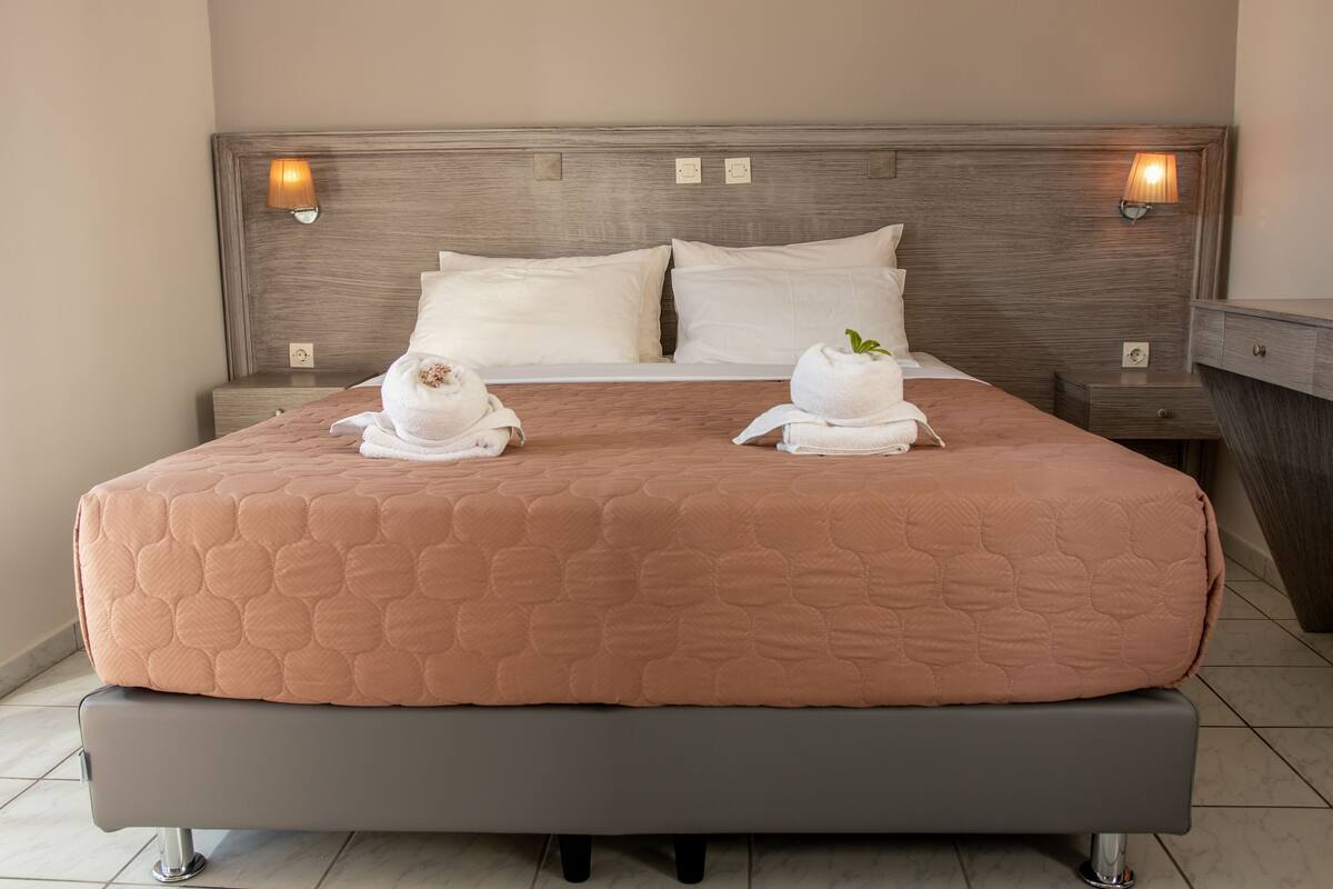 Una cama bien hecha con un edredón rosa, dos almohadas blancas y dos toallas, con una planta de menta encima. Hay dos lámparas de noche a cada lado de la cama.