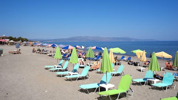 Una spiaggia con file di lettini e ombrelloni, cielo sereno e montagne sullo sfondo.