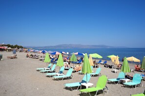 A beach with rows of sun loungers and umbrellas, a clear sky, and distant mountains. - Anna Katerina Apartments (Platanias)