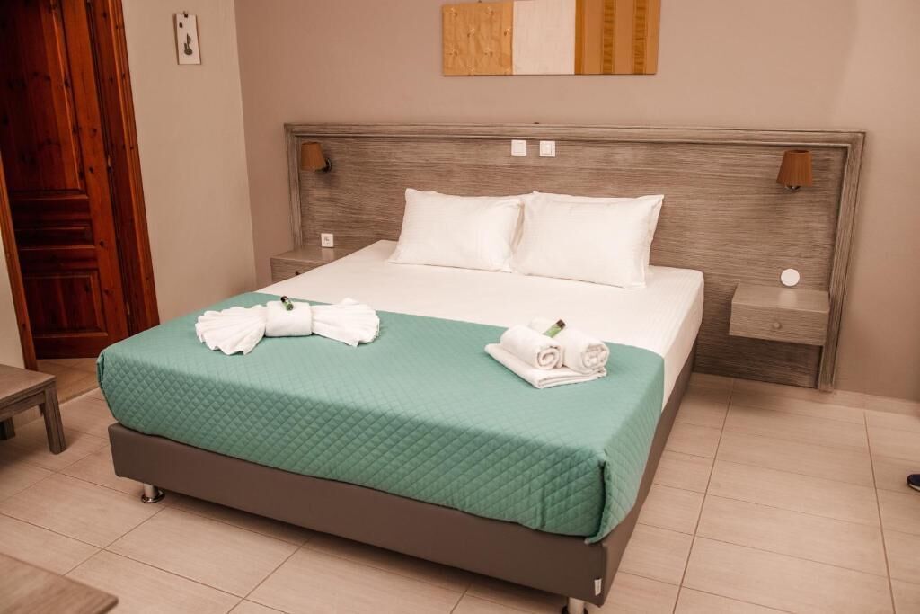 Una cama bien hecha con colcha verde y almohadas blancas, un cabecero de madera y una mesita de noche con una lámpara.