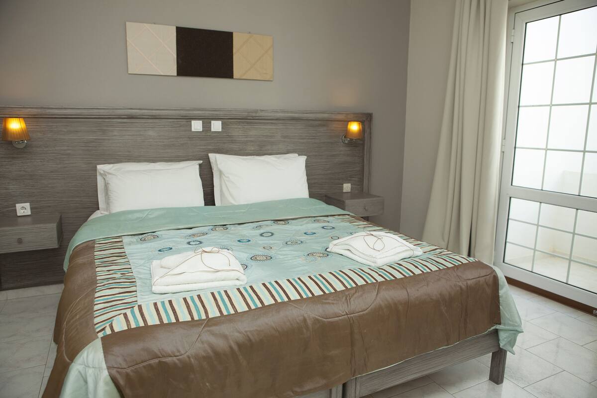 Una cama bien hecha con cabecera marrón, dos lámparas de noche y un ventanal con cortinas.