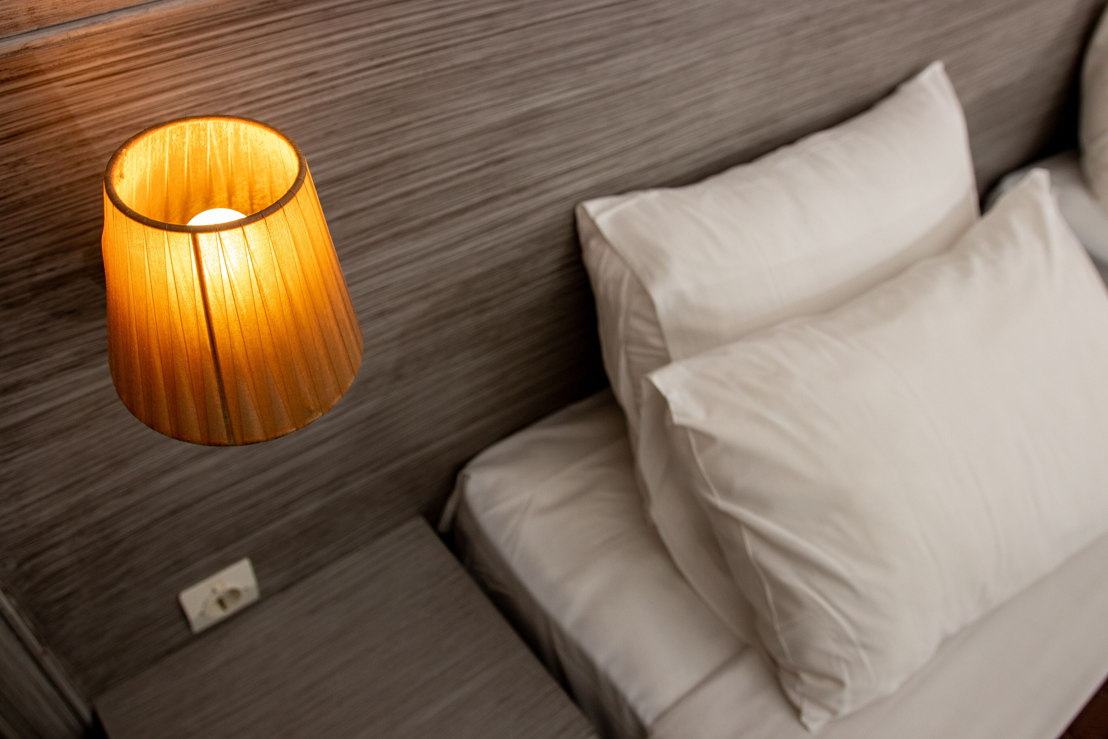 A modern room with a wooden headboard, a white bed with two pillows, and a cylindrical wall-mounted lamp.