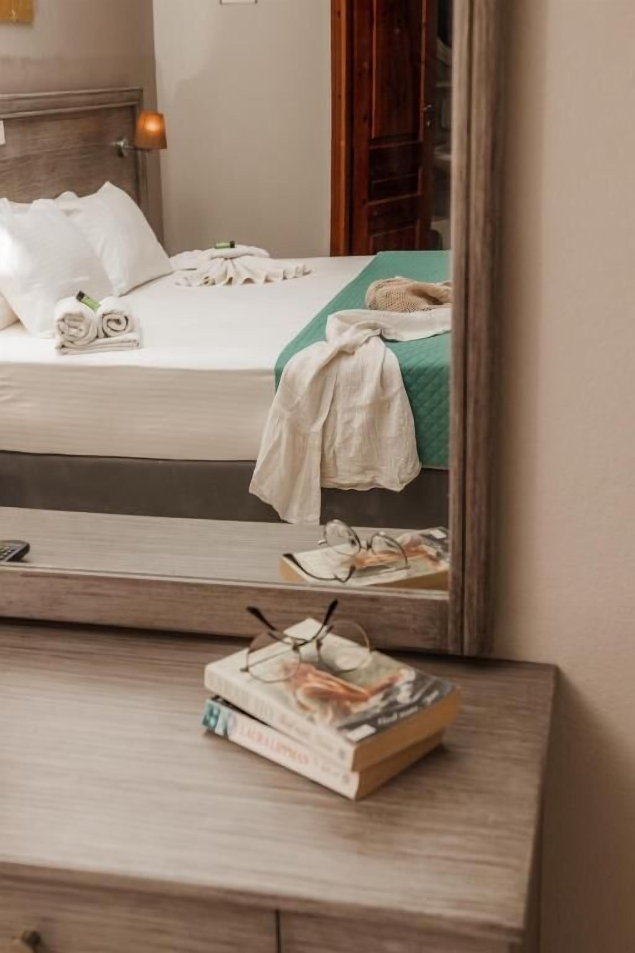 A neatly made bed with white linens, a green blanket, and a white towel. A bedside table holds a book and a pair of glasses.