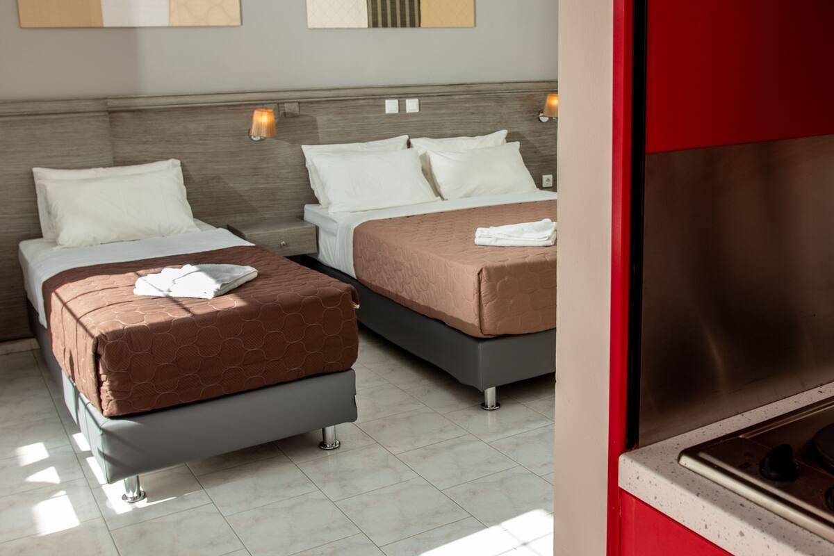 Habitación de hotel con dos camas, una pared roja, techo blanco y una zona de cocina.