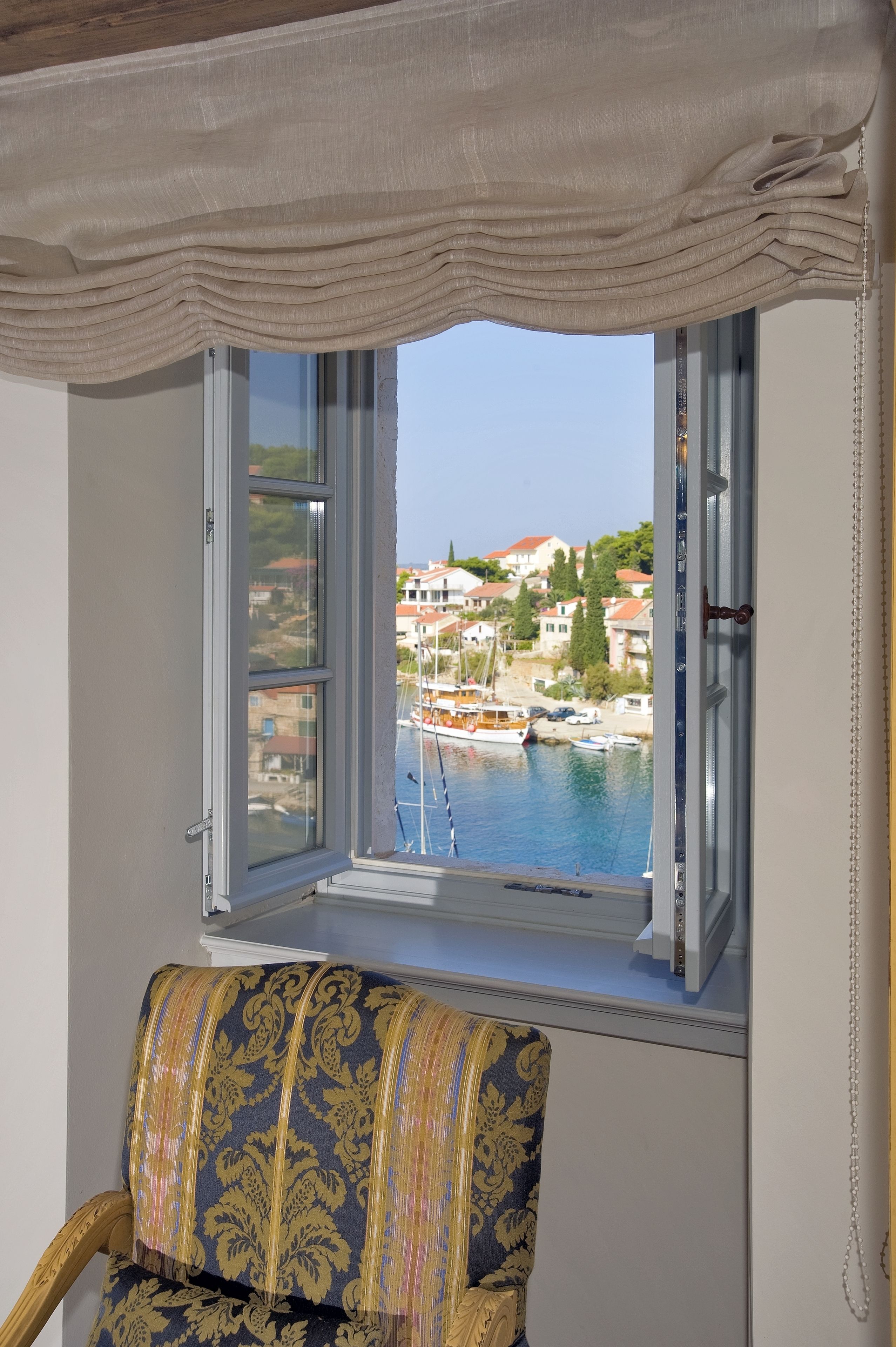 Une pièce avec vue sur un port, une fenêtre avec des rideaux et un fauteuil à motifs.