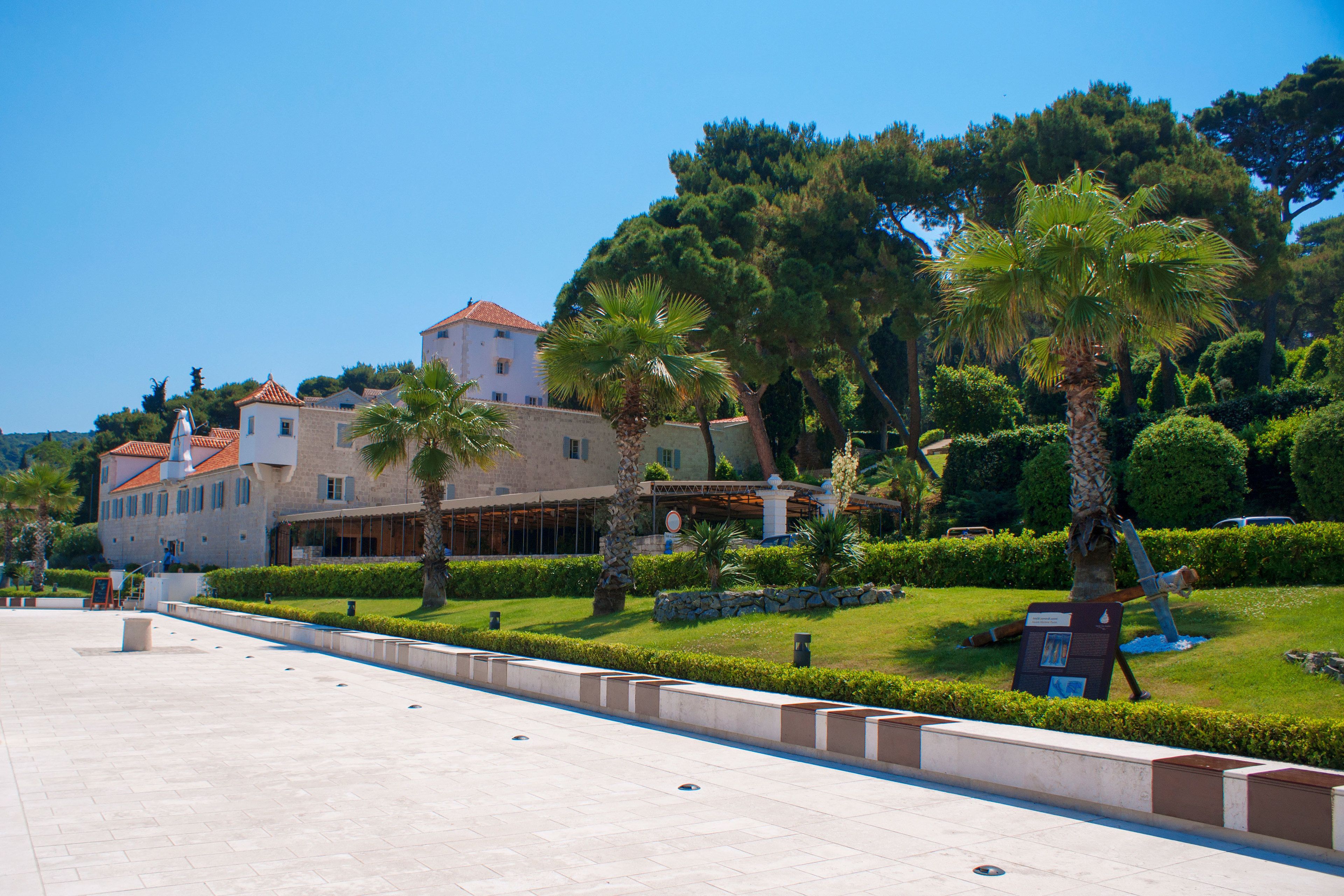 A spacious courtyard with a large building featuring a red-tiled roof, surrounded by well-maintained greenery and palm trees.