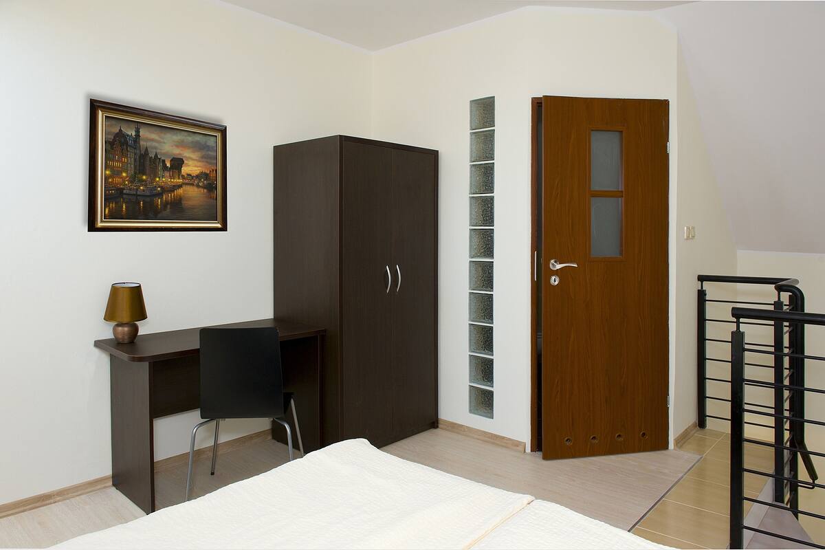 Un dormitorio con cama, escritorio con silla, armario, puerta con panel de vidrio y un cuadro en la pared.