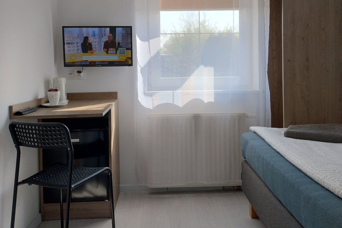 Una habitación pequeña con una cama, un escritorio y una silla. Hay una ventana con cortinas, un radiador y un televisor en la pared.