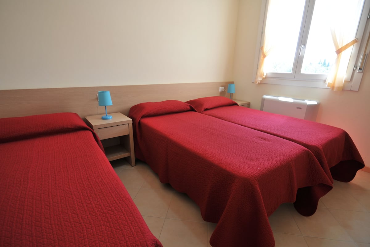 A room with two single beds, each with a red bedspread, a nightstand with a blue lamp, and a window with curtains.