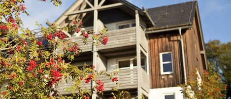 A wooden house with a balcony and a tree with red berries in the foreground.